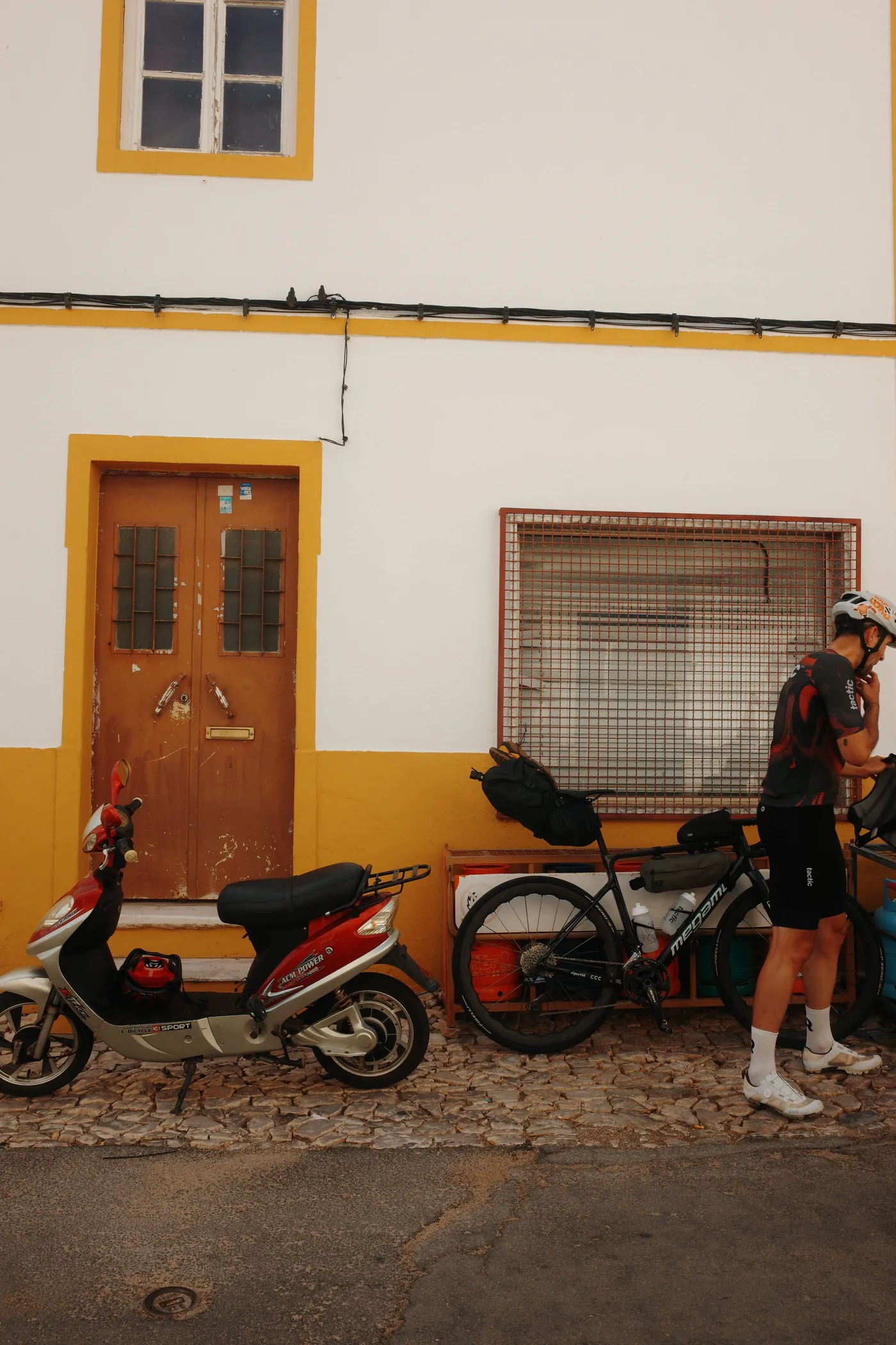 A loaded road bike and a scooter parked against a yellow-and-white house on a cobbled street