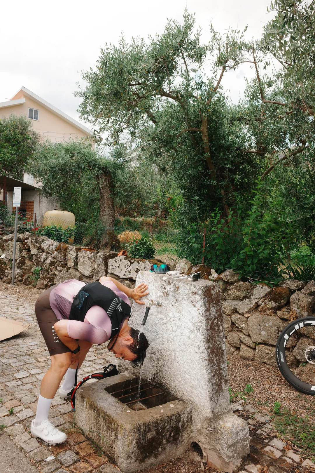 A cyclist bent over a stone village fountain with water running over the back of his head